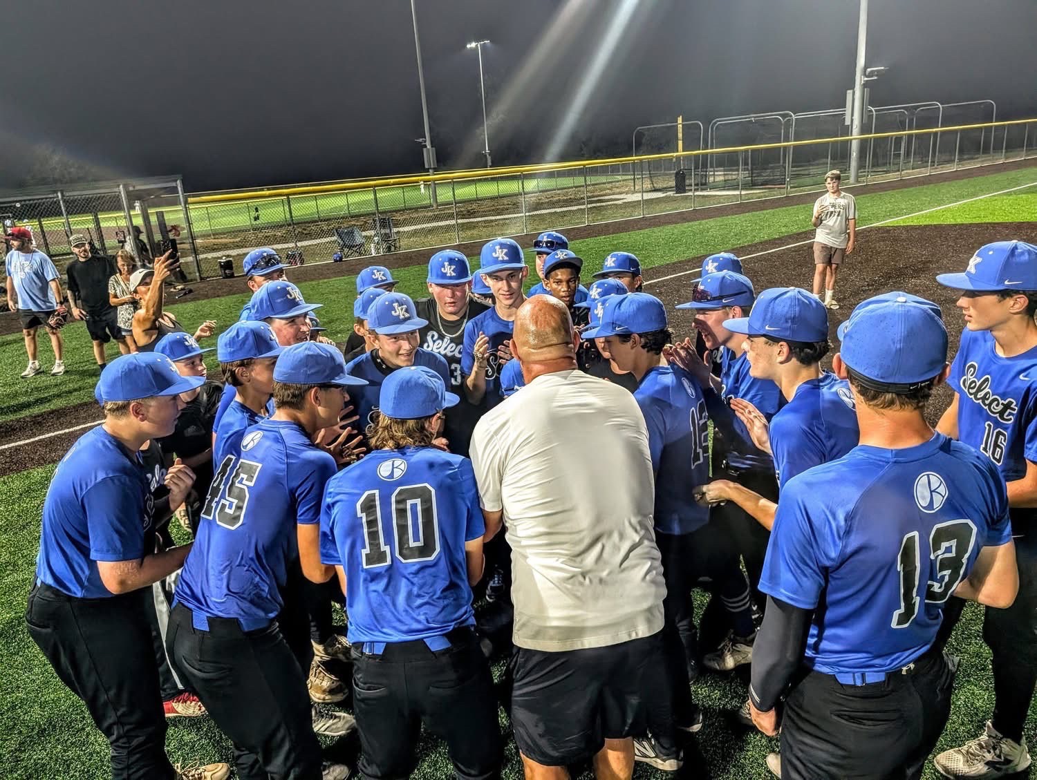 JK Select Missouri players in blue caps and jerseys gathered in a huddle around their coach under the stadium lights
